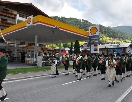 Eine Blaskapelle in traditionellen grünen Uniformen spielt Instrumente und geht an einer Shell-Tankstelle vorbei, mit Bergen im Hintergrund.