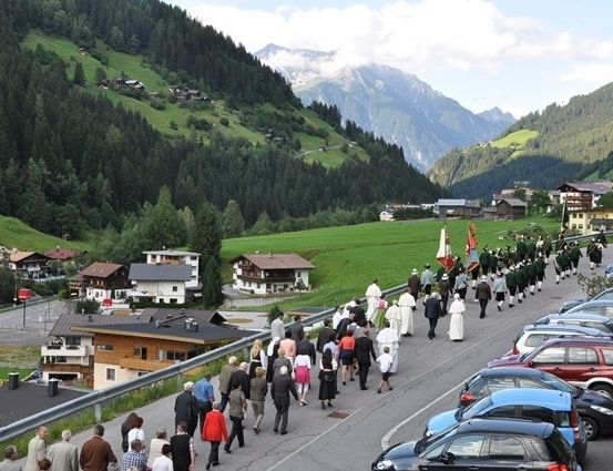 Eine Prozession von Menschen, einschließlich Priestern, geht auf einer Bergstraße, an der geparkte Autos stehen. Die Straße wird von Häusern gesäumt, und im Hintergrund befinden sich Berge.