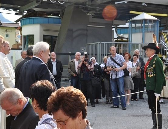 Eine Gruppe von Menschen, darunter ein Mann in einer grünen Uniform, versammelt sich um einen Priester in einer weißen Robe. Sie stehen vor einem Gebäude mit einer Fahne und einem Metallzaun.