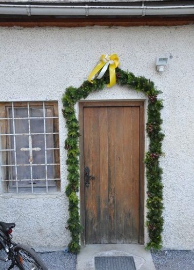Eine Holztür mit einem Kranz aus Grünpflanzen und einer gelben Schleife befindet sich an der Seite eines Gebäudes. Ein Fahrrad ist davor geparkt.