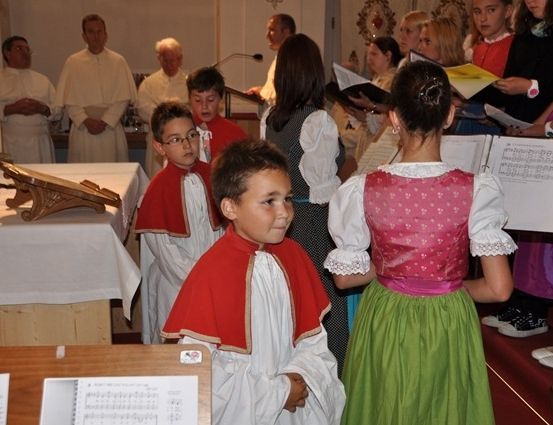 Eine Gruppe von Kindern in traditioneller Kleidung steht in einer Kirche, möglicherweise als Teil eines Chores. Hinter ihnen sind auch Erwachsene anwesend, einige halten Bücher und singen.