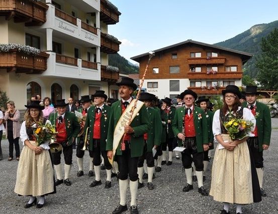 Eine Gruppe von Menschen in traditioneller Kleidung, die in einem Umzug vor einem Gebäude mit Balkonen marschiert. Einige halten Musikinstrumente und Blumen.