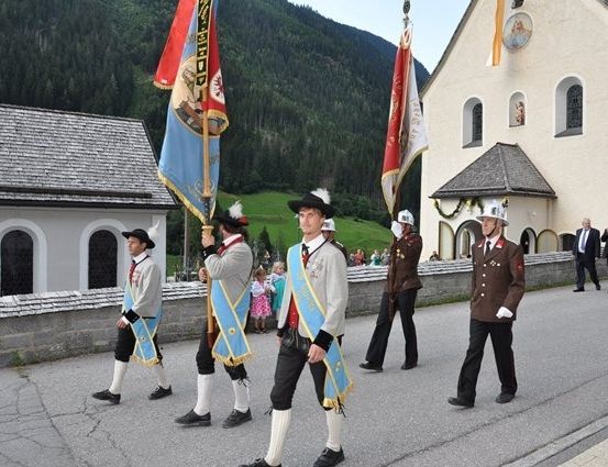 Eine Gruppe von Männern in traditionellen Uniformen marschiert vor einer Kirche mit Flaggen und Zuschauern.