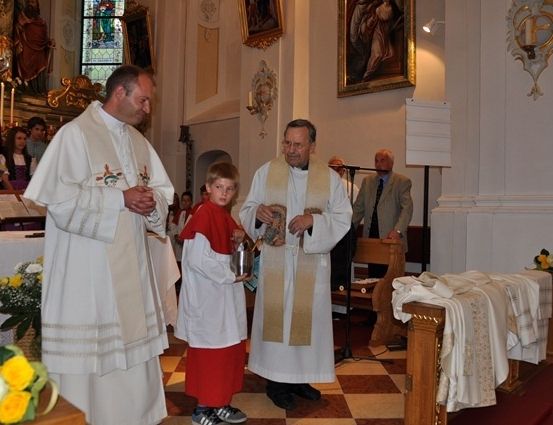 Zwei Priester stehen in einer Kirche, einer hält einen Kelch, während ein junger Junge in roten und weißen Gewändern einen kleineren Kelch hält. Dahinter sitzen eine Gruppe von Menschen.