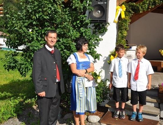 Ein Mann und eine Frau in traditioneller Kleidung stehen mit zwei Jungen vor einem geschmückten Gebäude. Der Mann trägt einen Anzug und eine Krawatte, und die Frau trägt ein blaues Kleid. Die Jungen sind in weißen Hemden und Krawatten.