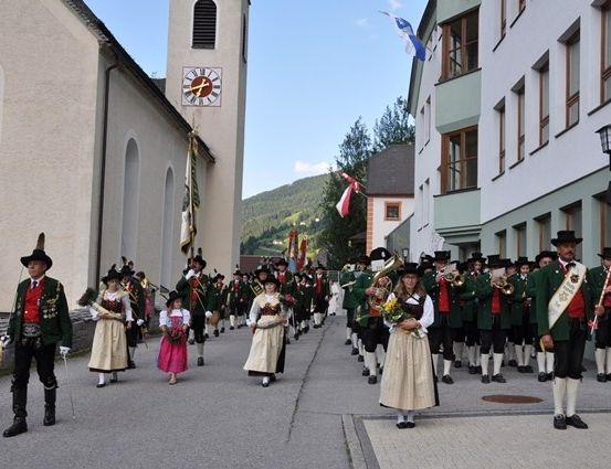 Eine Gruppe von Menschen in traditioneller Kleidung marschiert eine Straße hinunter, mit Gebäuden und einem Uhrturm im Hintergrund.