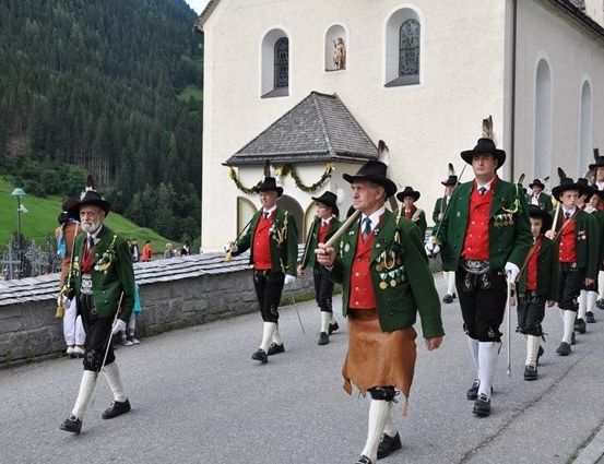 Eine Gruppe von Männern in traditionellen grünen und roten Uniformen, die Stöcke tragen, in einer Reihe auf einer Straße vor einer Kirche marschieren.