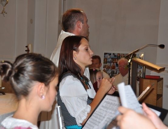 Eine Gruppe von Menschen versammelt sich in einer Kirche, wobei eine Frau in traditioneller Kleidung ein Notenblatt hält. Ein Priester steht hinter ihr, und andere sind im Gebet vertieft.