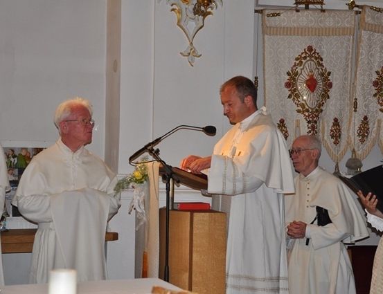 Ein Priester liest von einem Podium während eines Gottesdienstes, mit mehreren anderen Geistlichen und einer Frau in der Nähe.