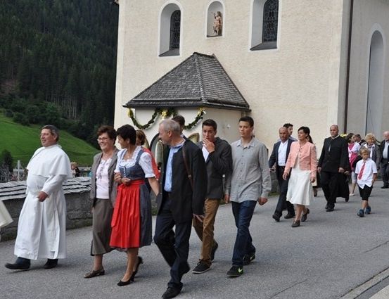 Eine Gruppe von Menschen, einschließlich eines Priesters in einer weißen Robe, geht vor einer Kirche in einem Gebirgsgebiet.