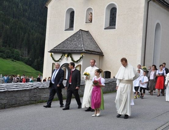 Eine Gruppe von Menschen, darunter Priester und ein Mädchen in traditioneller Kleidung, gehen auf einer Straße vor einer Kirche mit einem grünen Hügel im Hintergrund.