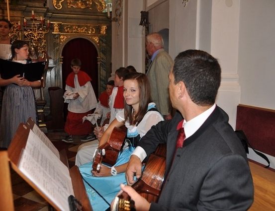 Eine Gruppe von Menschen versammelt sich in einer Kirche, wobei ein Mann und eine Frau Gitarre spielen und ein Mädchen aus einem Buch liest.