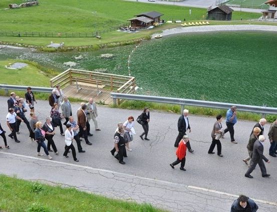 Eine Gruppe von Menschen geht auf einer Straße in der Nähe eines Sees, mit einer Holzbrücke und Häusern im Hintergrund.