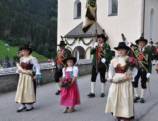 Eine Gruppe von Menschen in traditionellen Kostümen, einschließlich Kinder, gehen in einer Parade vor einer Kirche mit einer Fahne.