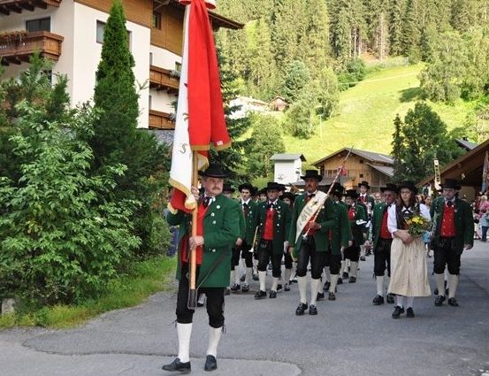 Eine Gruppe von Menschen in traditionellen grünen Uniformen und Hüten marschiert bei einer Parade mit einer Fahne vor einem Gebäude und Bäumen.