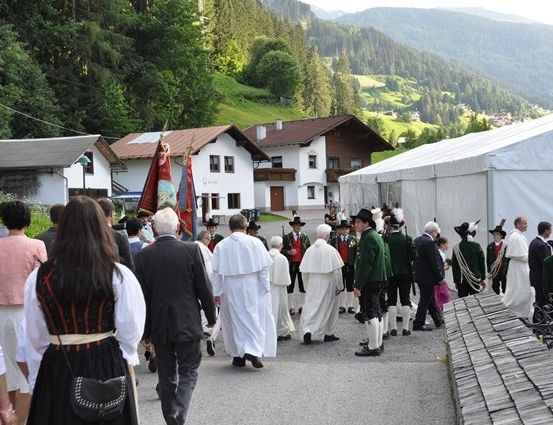 Eine Gruppe von Menschen in traditioneller Kleidung, einschließlich Geistlicher, marschiert in einer Prozession in einem Bergdorf mit Häusern und einem Zelt im Hintergrund.