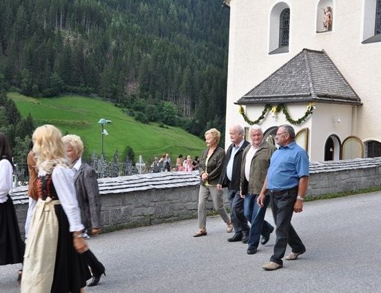 Eine Gruppe von Menschen, einige in traditioneller Kleidung, geht an einer Kirche mit einem Kreuz und Grünpflanzen im Hintergrund vorbei.