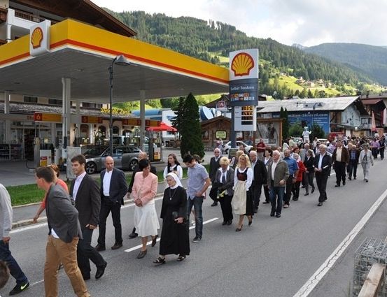 Eine Gruppe von Menschen, darunter eine Nonne, geht vor einer Shell-Tankstelle mit Bergen im Hintergrund die Straße entlang.