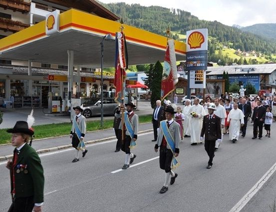 Ein Zug von Personen in traditioneller Kleidung, einschließlich eines jungen Jungen in Grün, marschiert die Straße hinunter, mit einer Shell-Tankstelle im Hintergrund.