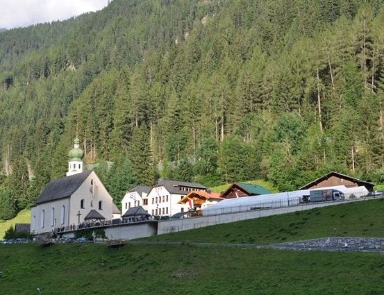 Ein kleines Dorf mit einer Kirche und mehreren Häusern in einem grünen Tal, umgeben von hohen Bäumen.