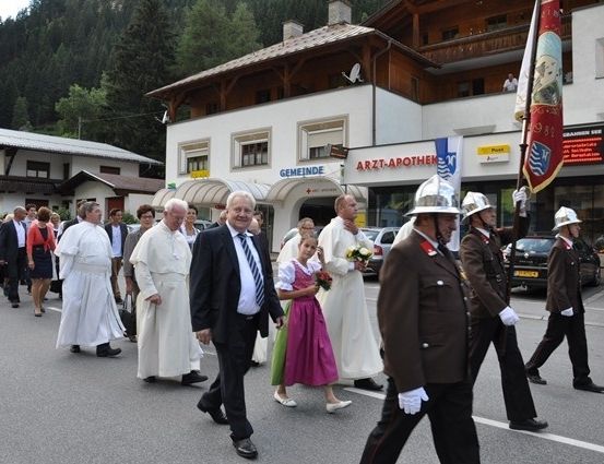 Eine Gruppe von Menschen, einschließlich Priester, gehen in einem Umzug auf einer Straße mit Gebäuden und Bergen im Hintergrund.