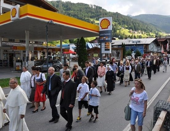 Eine Gruppe von Menschen, darunter Geistliche, geht vor einer Shell-Tankstelle mit Bergen im Hintergrund die Straße entlang.
