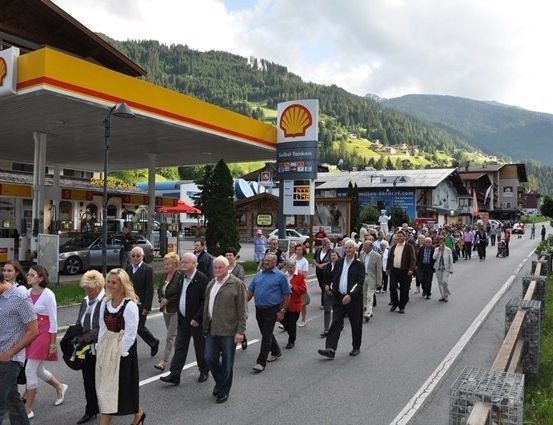 Eine Gruppe von Menschen geht eine Straße entlang, die von Geschäften und Tankstellen gesäumt ist, mit Bergen im Hintergrund.