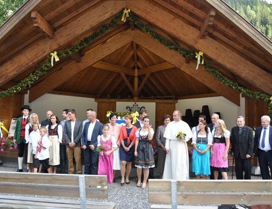 Eine Gruppe von Menschen, einschließlich eines Priesters, steht vor einer hölzernen Kapelle mit dekorativen Kränzen und Bändern. Sie sind in traditioneller Kleidung gekleidet.