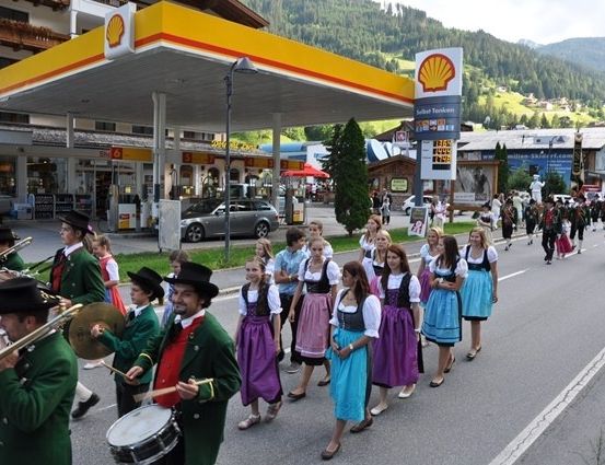 Eine Blaskapelle in traditioneller Kleidung, die Instrumente spielt, führt einen Umzug die Straße entlang, mit einer Shell-Tankstelle im Hintergrund.