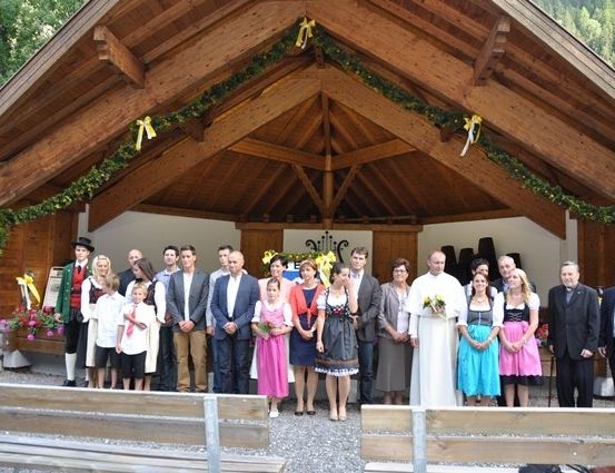 Eine Gruppe von Menschen, darunter Erwachsene und Kinder, versammelt sich vor einer hölzernen Kapelle mit festlicher Atmosphäre, möglicherweise für eine Hochzeitszeremonie.