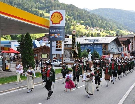 Eine Gruppe von Menschen in traditioneller Kleidung marschiert eine Straße hinunter, mit einer Shell-Tankstelle auf der linken Seite und Bergen im Hintergrund.