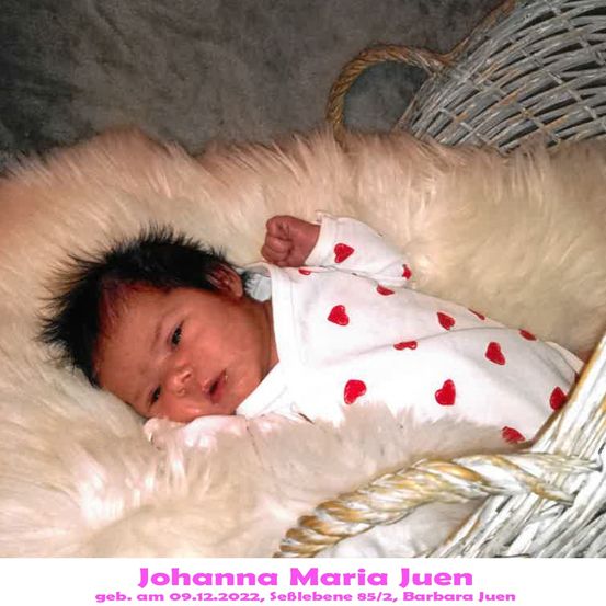 Ein Babymädchen namens Johanna Maria Juen, geboren am 9. Dezember 2022, liegt auf einer weißen Pelzdecke in einem Korb. Sie trägt ein weißes Strampler mit roten Herzdesigns.