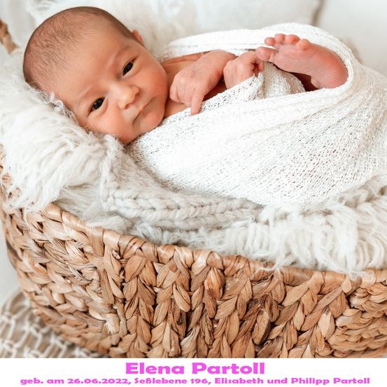 Ein neugeborenes Baby, in eine weiße Decke gewickelt, liegt in einem Korb mit weißer Fellfütterung und lächelt direkt in die Kamera. Das Baby heißt Elena Partoll, geboren am 26. Juni 2022.