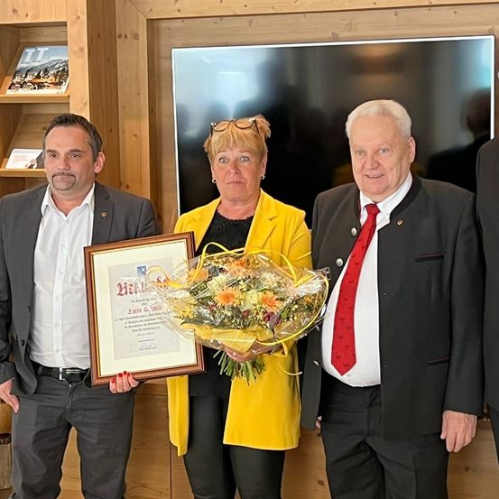 Drei Personen stehen vor einer Holzwand, eine hält ein gerahmtes Zertifikat und einen Blumenstrauß, während die anderen lächeln und in die Kamera schauen.