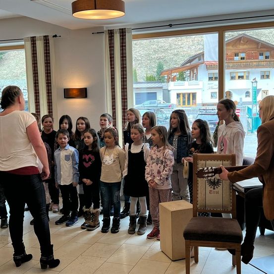 Eine Gruppe von Kindern und zwei Erwachsenen steht in einem Raum und singt, während eine Person die Gitarre spielt. Der Raum hat große Fenster und einen Blick auf einen Berg und Häuser.