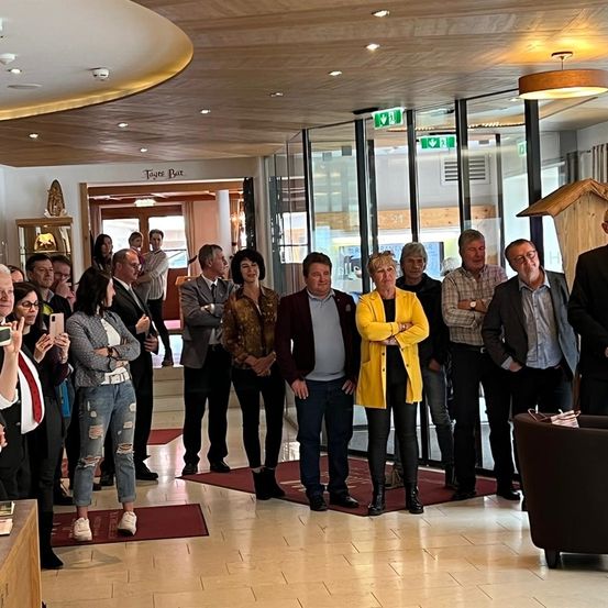 Eine Gruppe von formell gekleideten Menschen steht in einer Lobby, einige machen Fotos. Die Lobby hat ein modernes Design mit Glaswänden und einer Holzstruktur.