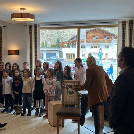 Eine Gruppe von Kindern und zwei Erwachsenen steht in einem Raum und singt, während eine Frau ein Buch in der Hand hält. Ein Stuhl mit einem Logo steht vor ihnen.