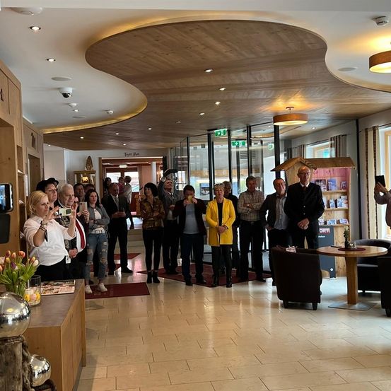 Eine Gruppe von Menschen in formeller Kleidung steht in einer modernen Lobby, einige machen Fotos mit ihren Handys. Der Raum hat Holzböden, große Fenster und eine Holzdecke.