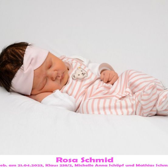 Ein neugeborenes Mädchen namens Rosa Schmid, geboren am 21. April 2023, schläft friedlich auf einem weißen Kissen. Sie trägt ein rosa-weiß gestreiftes Outfit mit einem Teddybär-Patch.