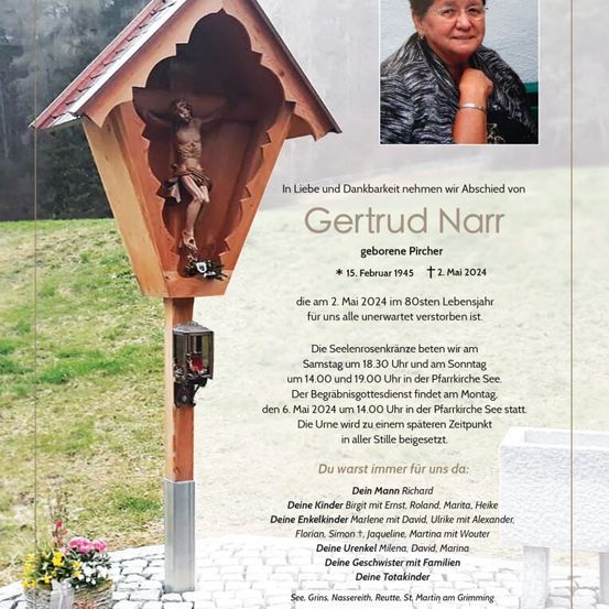 Ein Gedenkschild mit einem Bild einer Frau, Gertrude Narr, geboren am 15. Februar 1945 und verstorben am 2. Mai 2024. Das Schild zeigt ein Holzkreuz und Blumen. Der Hintergrund ist eine Rasenfläche mit einer Holzstruktur.
