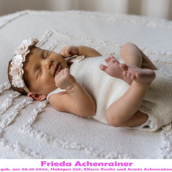 Ein neugeborenes Babymädchen liegt auf einer weißen Decke und trägt ein weißes gestricktes Outfit und eine Blumenhaarschleife. Sie lächelt und scheint friedlich zu schlafen. Das Bild hat einen Wasserzeichen mit dem Text 'Frieda Achenrainer' und dem Datum '08.10.2024'.