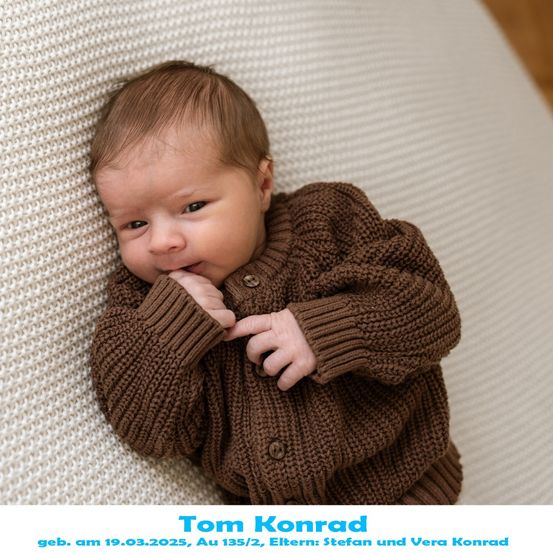 Ein Babyjunge namens Tom Konrad, geboren am 19.03.2025, liegt auf einer weißen gestrickten Decke mit den Händen am Kinn. Er trägt einen braunen gestrickten Pullover.
