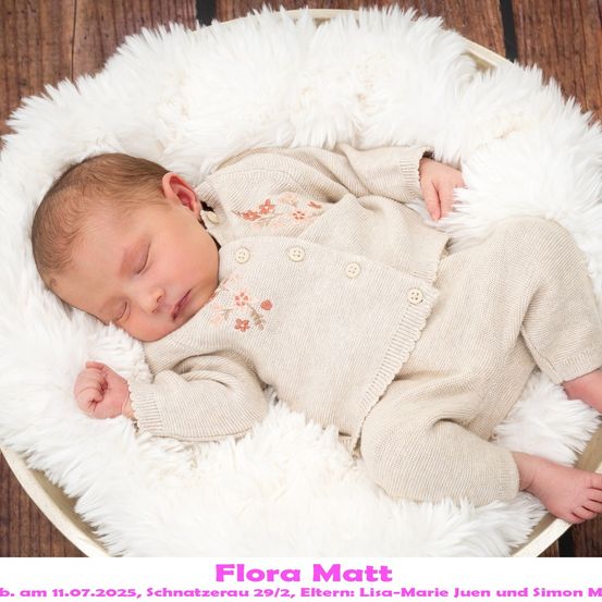 Ein schlafendes Babymädchen in einem beigen Outfit mit Blumenstickerei, das in einem runden Korb auf einer weißen Pelzdecke liegt. Der Name des Babys ist Flora Matt, geboren am 11. Juli 2025.