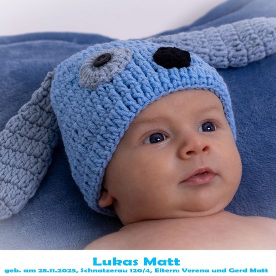 Ein Babyjunge, der eine blaue gestrickte Mütze mit Hundegesichtsdesign trägt, liegt auf einer blauen Decke. Der Babyjunge heißt Lukas Matt, geboren am 28. November 2025.