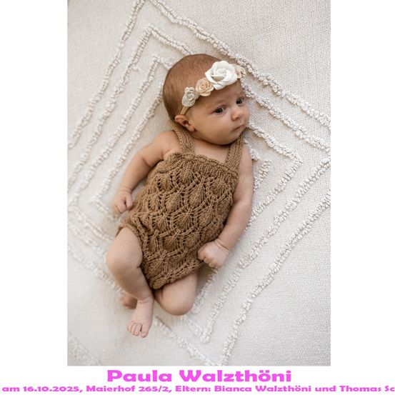 Ein Babymädchen liegt auf einer weißen Decke und trägt ein braunes gestricktes Kleid und eine weiße Blumenhaarband. Das Bild wurde am 16. Oktober 2025 in Maierhof 265/2 aufgenommen.