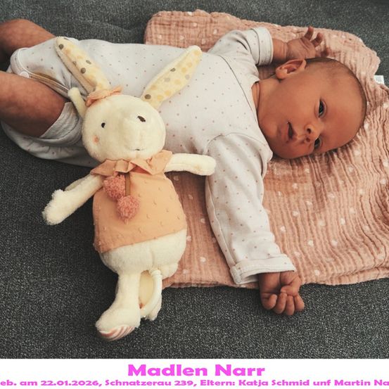 Ein Baby liegt auf einer rosa Decke mit einem Plüschhasen-Spielzeug, lächelt und schaut in die Kamera. Das Baby trägt ein weißes Langarm-Outfit mit Punkten. Das Bild hat einen Wasserzeichen-Text am unteren Rand.