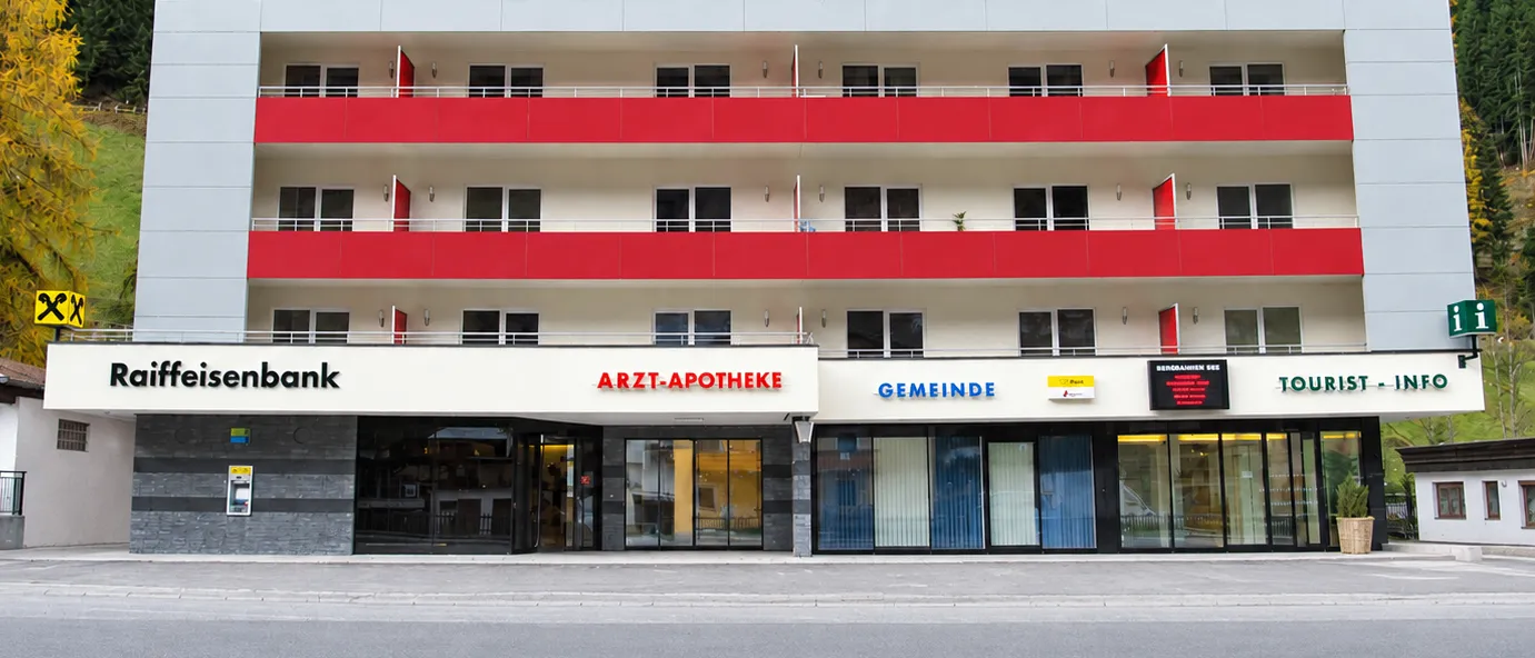 Ein modernes Gebäude mit roten Balkonen und weißen Wänden, mit einer Apotheke, einem Gemeindezentrum und einem Tourismusbüro im Erdgeschoss.