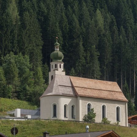 Eine kleine weiße Kirche mit einer grünen Kuppel und einem braunen Dach steht auf einem Hügel, umgeben von hohen Bäumen und Grün.