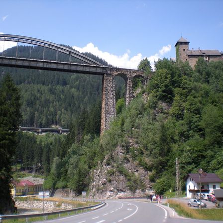 Eine große Steinbrücke mit einem Bogenbauwerk ist über einem steilen Berg gebaut, mit einer Burg auf der Spitze. Unter der Brücke ist eine Straße mit Autos und Häusern sichtbar.