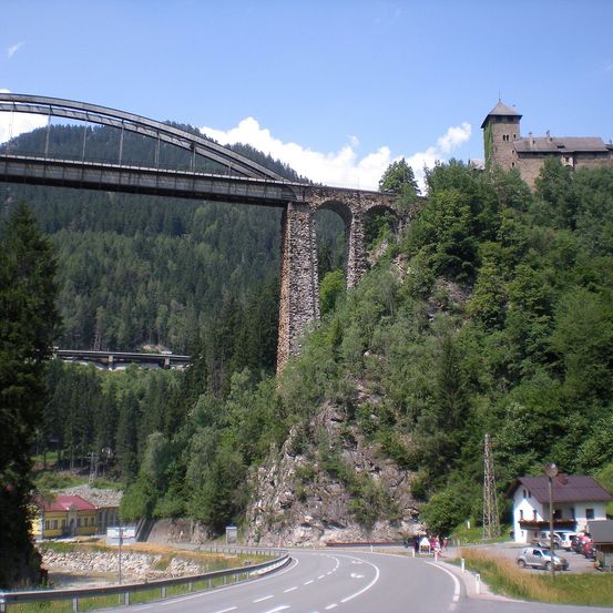 Eine große Steinbrücke mit einem Bogenbauwerk ist über einem steilen Berg gebaut, mit einer Burg auf der Spitze. Unter der Brücke ist eine Straße mit Autos und Häusern sichtbar.